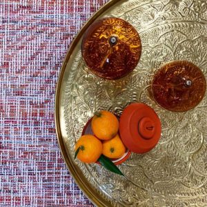Photographie d'un plateau doré Bazardeluxe sur un tapis rose. Sur le plateau il ya des lampes à huile en verre, une boite en carton orange Alice Deloule avec des clémentines